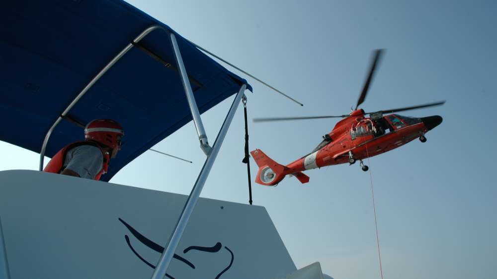 An orange helicopter hovers over a white boat.