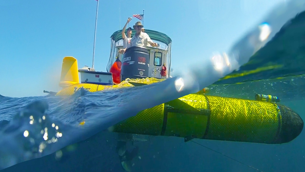 A yellow ocean glider floats on the ocean surface with people standing on a boat behind it.