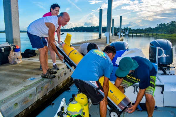 A group of people move a yellow torpedo-shaped piece of equipment from a boat dock
								onto a boat.