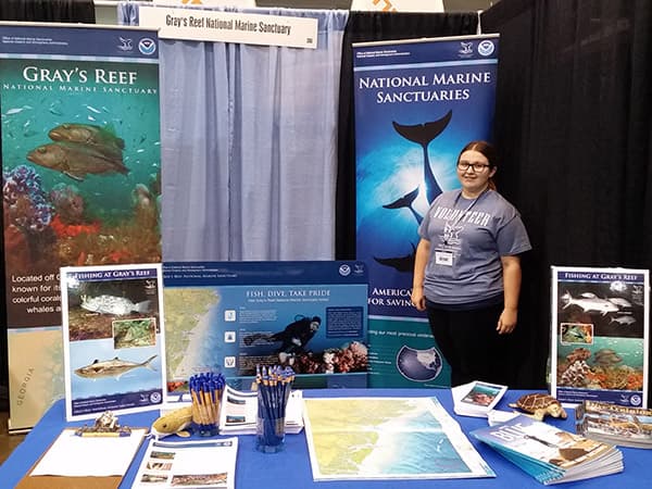 A person stands behind 6-foot table decorated with photos and banners of Gray's Reef National Marine Sanctuary, at the Savannah Boat Show