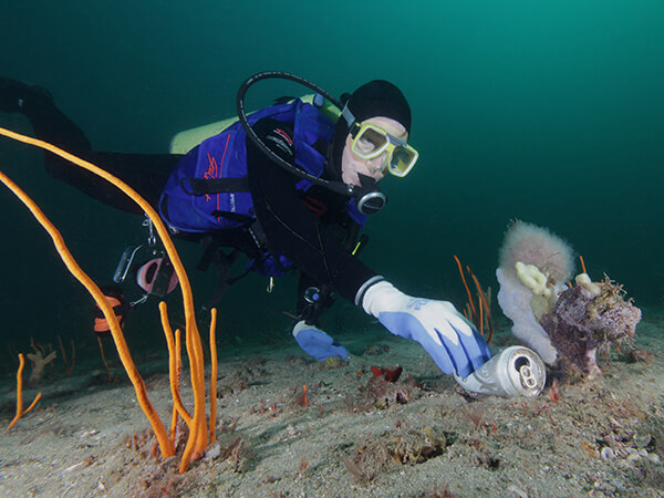 A scuba diver reaches to pick up an aluminum can on a sandy seafloor.