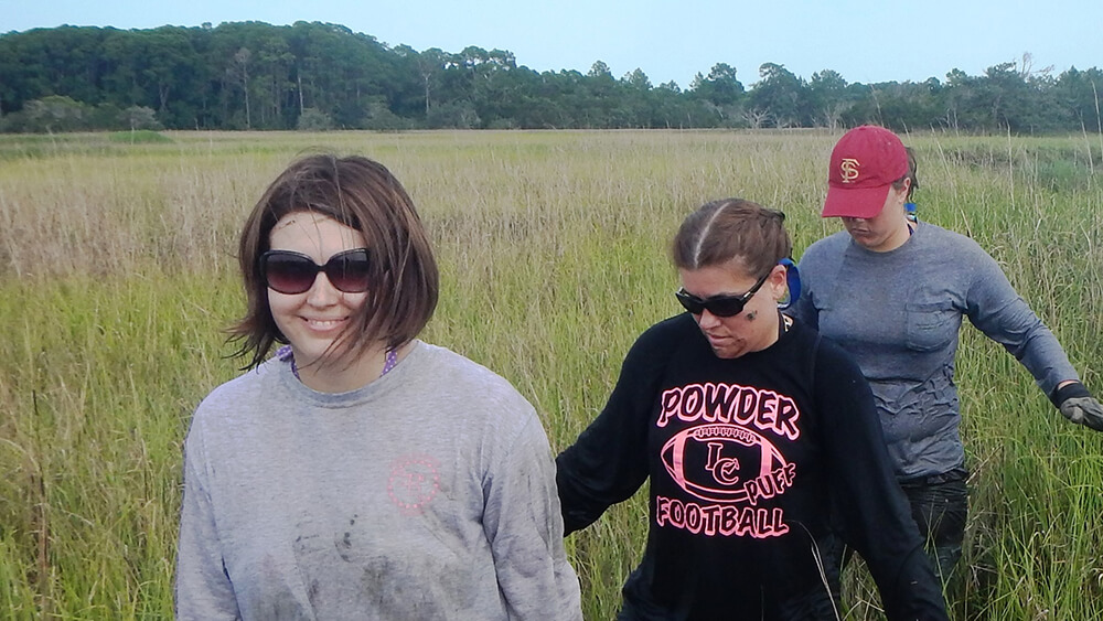 Three people walk through a salt marsh in a single file line.