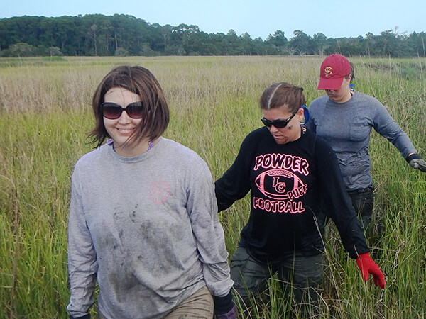 Three people walk through a salt marsh in a single file line.