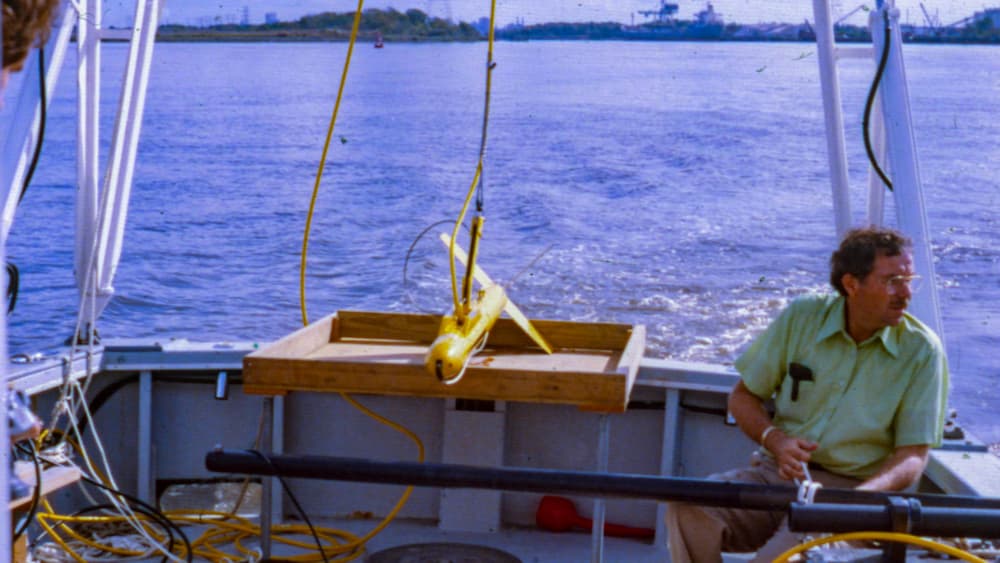 A person sits on a boat deck with a yellow torpedo-shaped piece of equipment that is used to map the ocean floor.