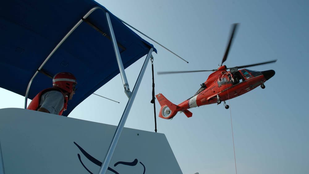 An orange helicopter hovers over a boat while a helmeted person looks on.