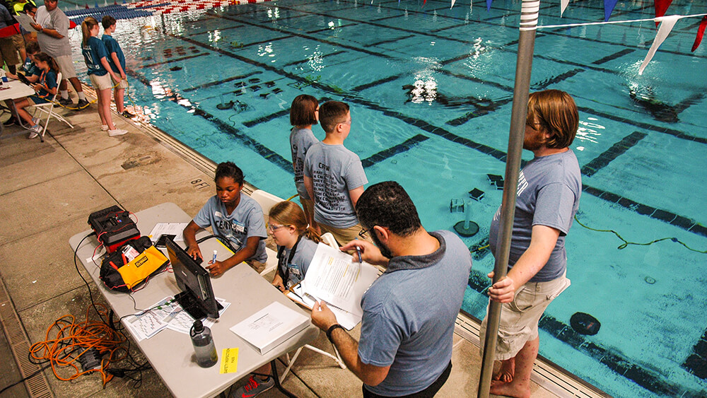 People stand next to an indoor pool with robotic equipment on folding tables.